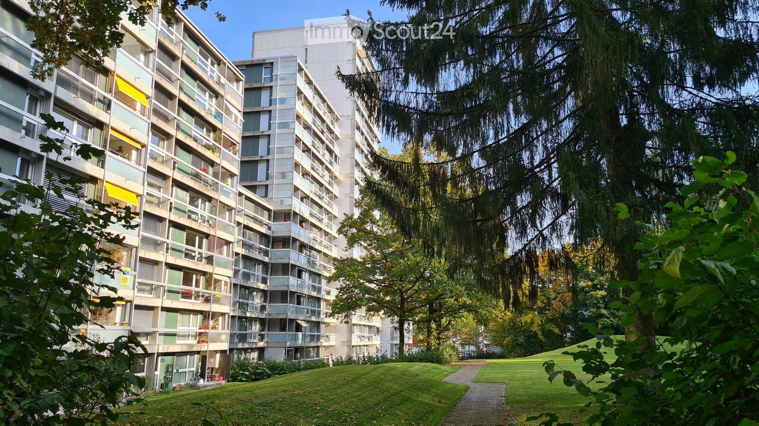 Two residential apartment buildings, each with multiple floors and balconies, surrounded by a grassy field and trees.