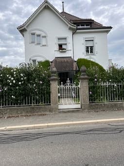 white house with a brown roof, white windows, small garden, large fence