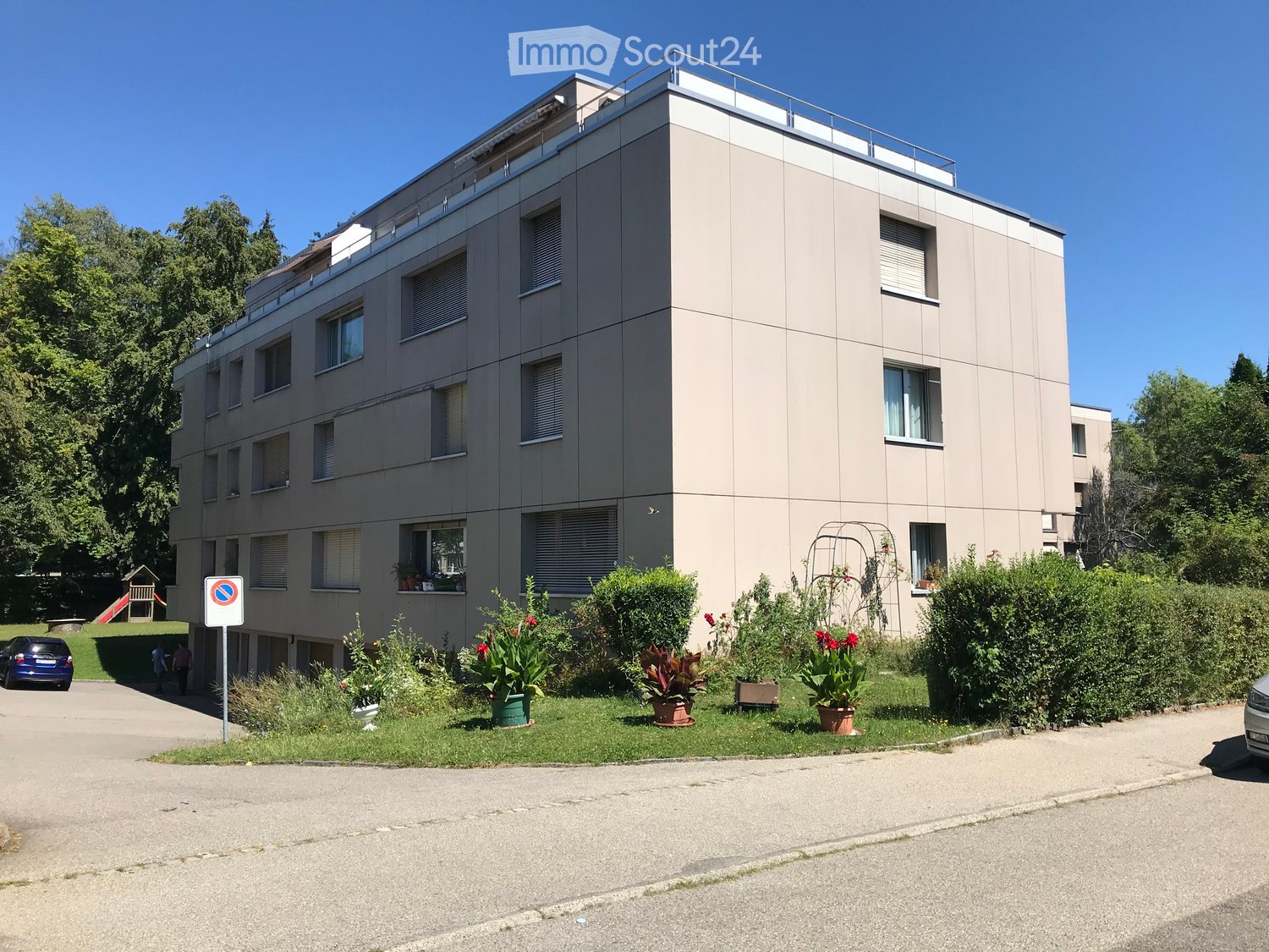Apartment building with 5 floors, large windows, plants in the foreground, green grass, fence, cars parked in the driveway