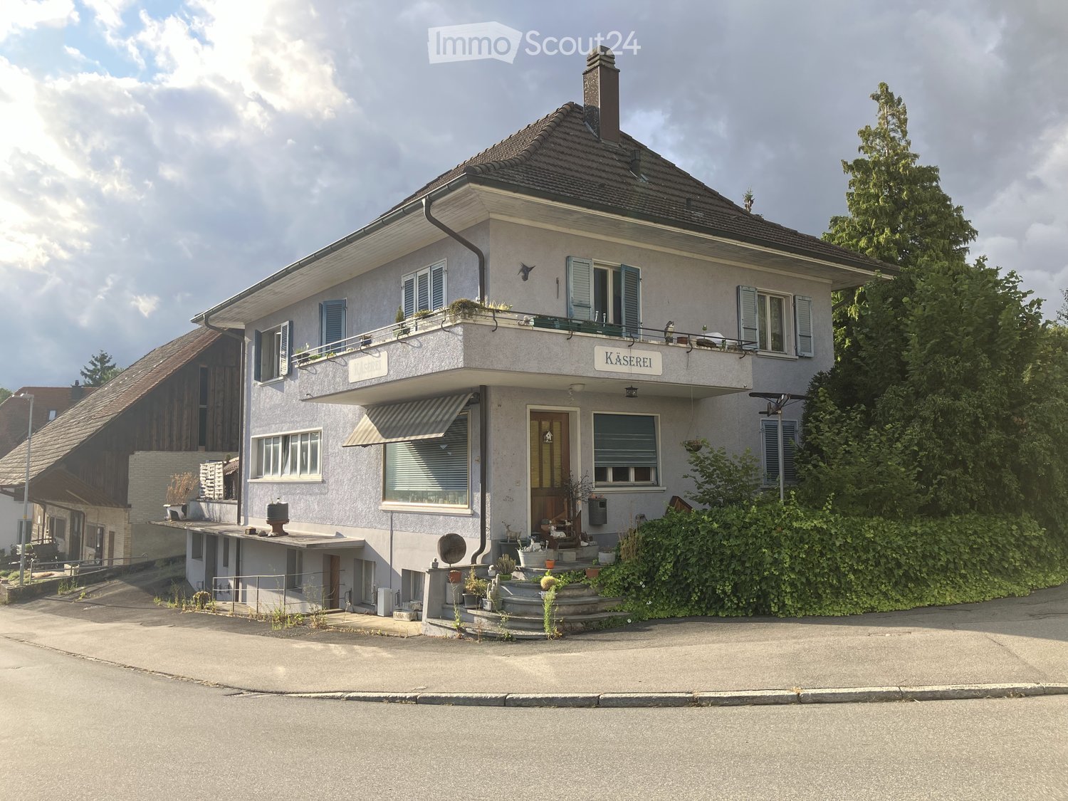 white stucco building, balcony, window awnings, plants and plants in front of the entrance, small garden, sign that reads Kaserei