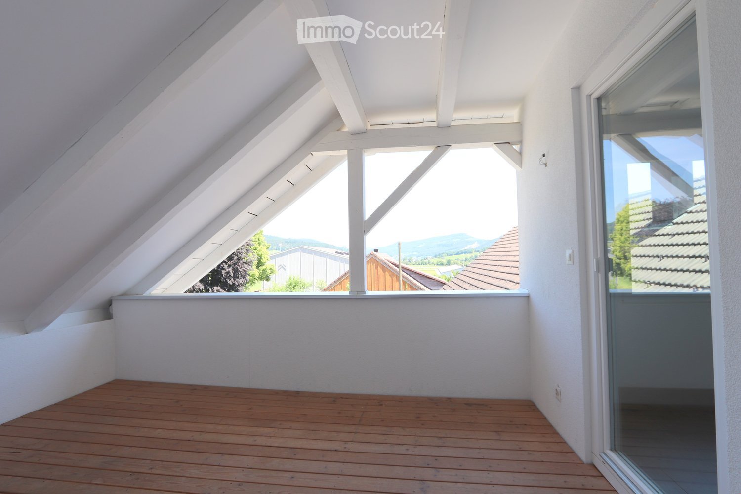 an empty attic room with wooden floor, white walls, beams, and windows with outside view
