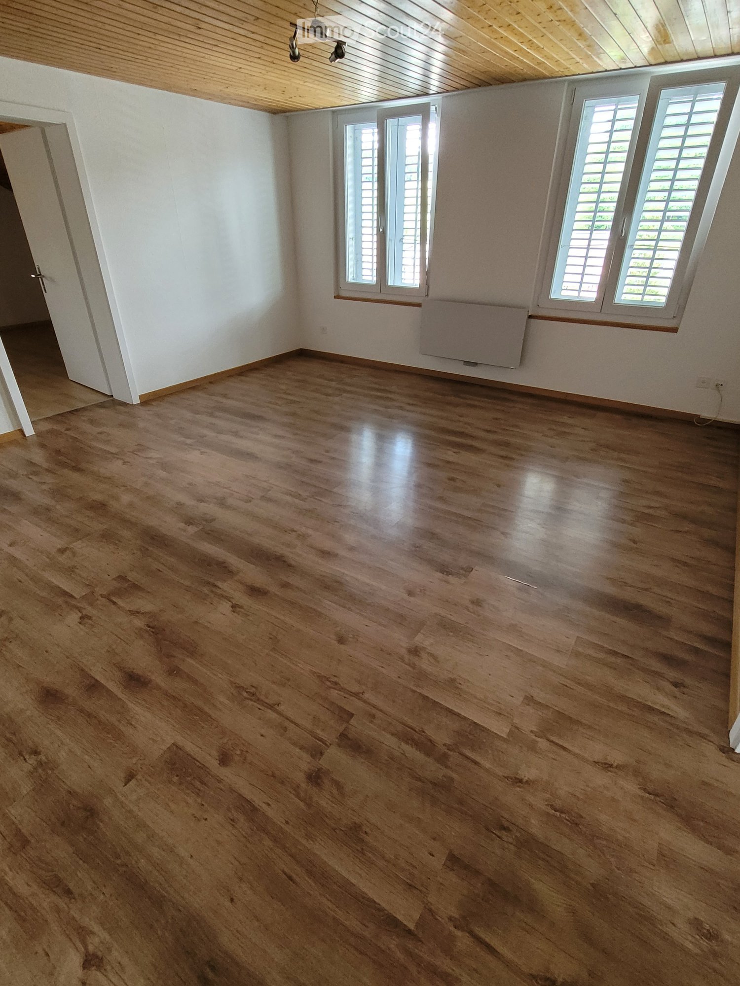 Empty room with wooden flooring, white walls, windows with shutters, and a ceiling lamp.