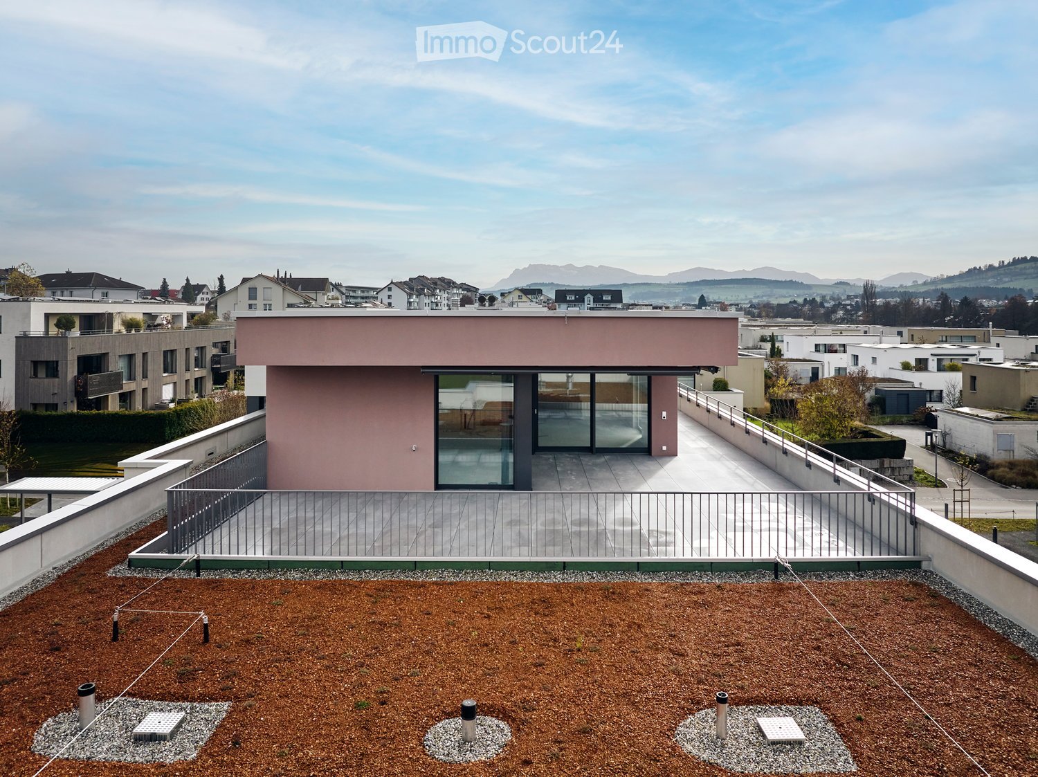 Rooftop building with a balcony and panoramic view, made of pink materials, multiple glass windows, surrounded by trees and other buildings