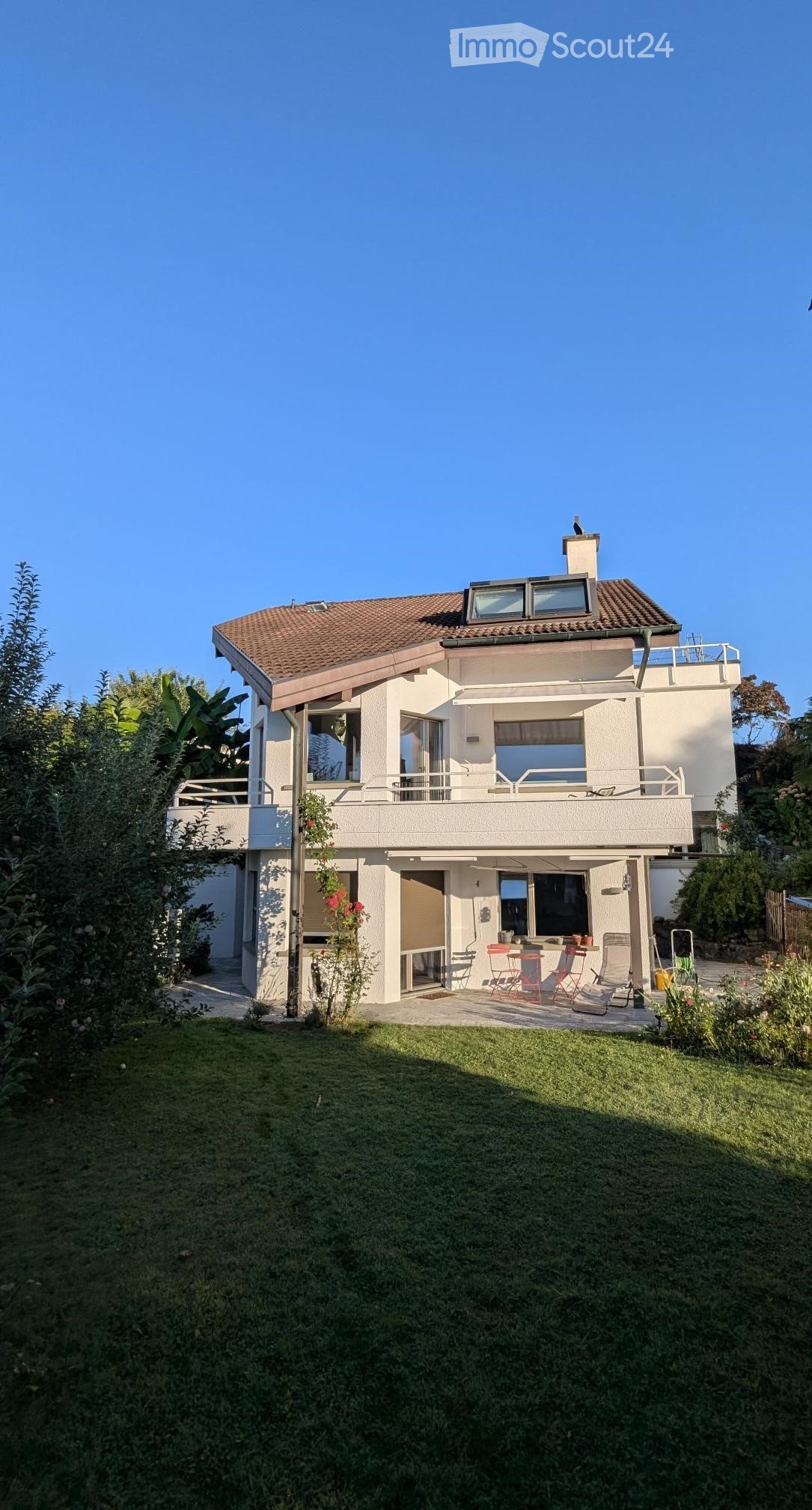 2 story house, brown roof, white walls, balcony, green lawn, garden with flowers, outdoor chairs and table