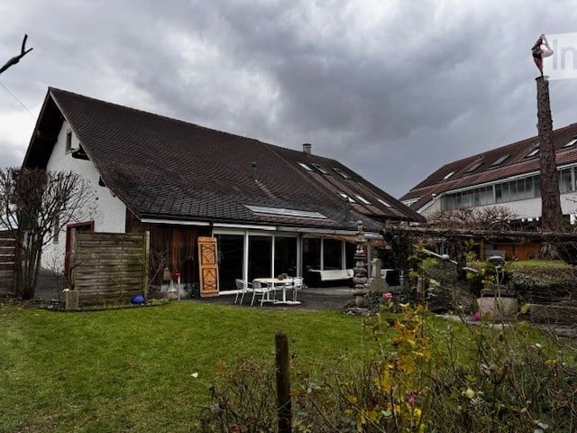 House with raised ground floor, brown roof, white walls, solar panels on roof, glass windows, wooden entrance doors, garden with grass and plants, white outdoor table and chairs, other building in the background