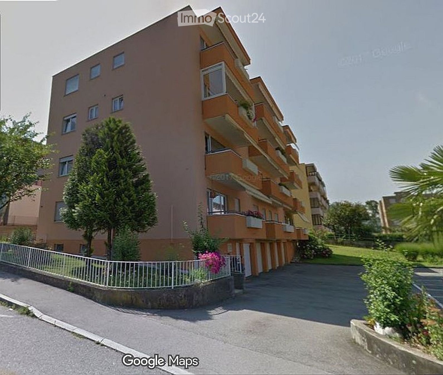 Apartment building, balconies, fenced garden, paved road, large green grass area, trees
