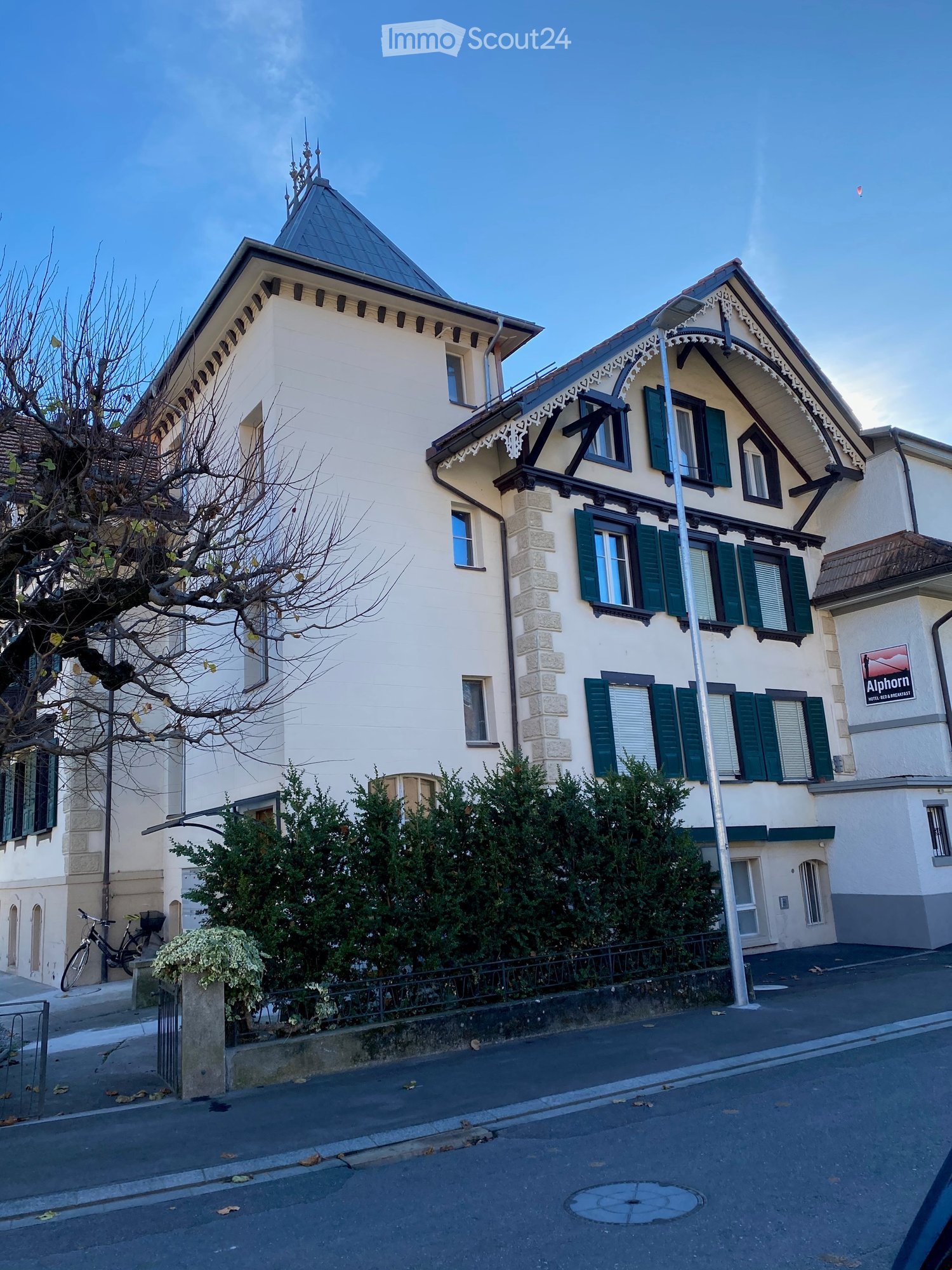 Multi-story white building with green shutters, triangular roof, stone walls, sign 'Alphorn', bicycle parked outside, street light.