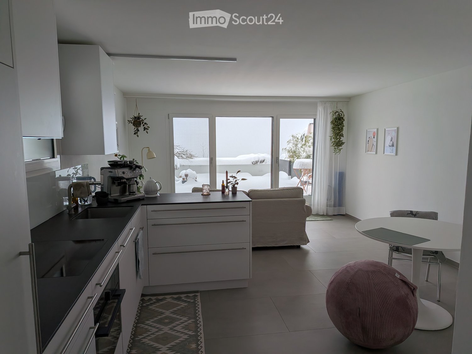 Modern kitchen with granite countertops, white cabinets, a kitchen island, and open layout to living room. Living room has sofa, rug, and a table with chairs. Sliding doors leading to balcony.