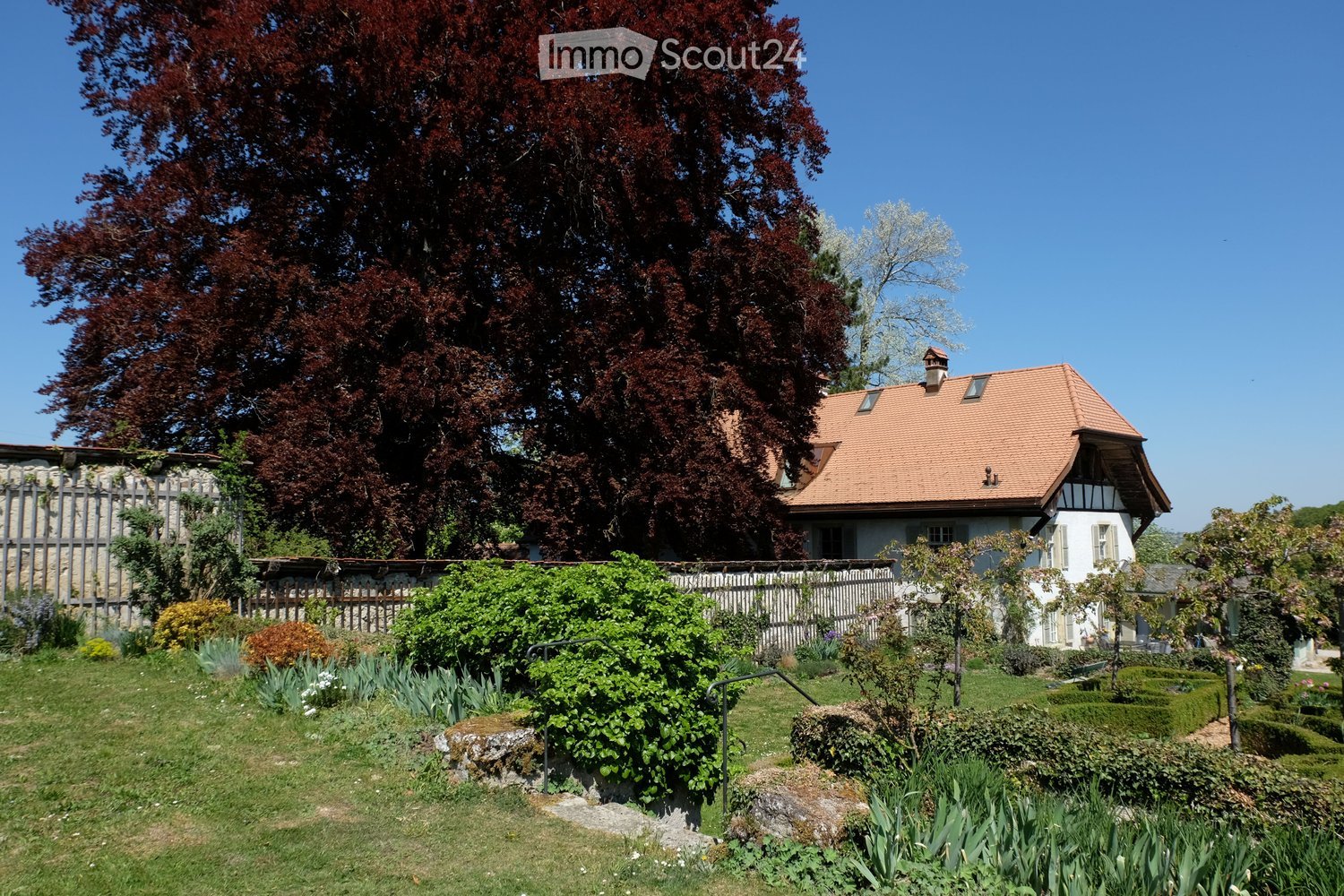 House with red roof and chimney, surrounded by green lawn and manicured bushes