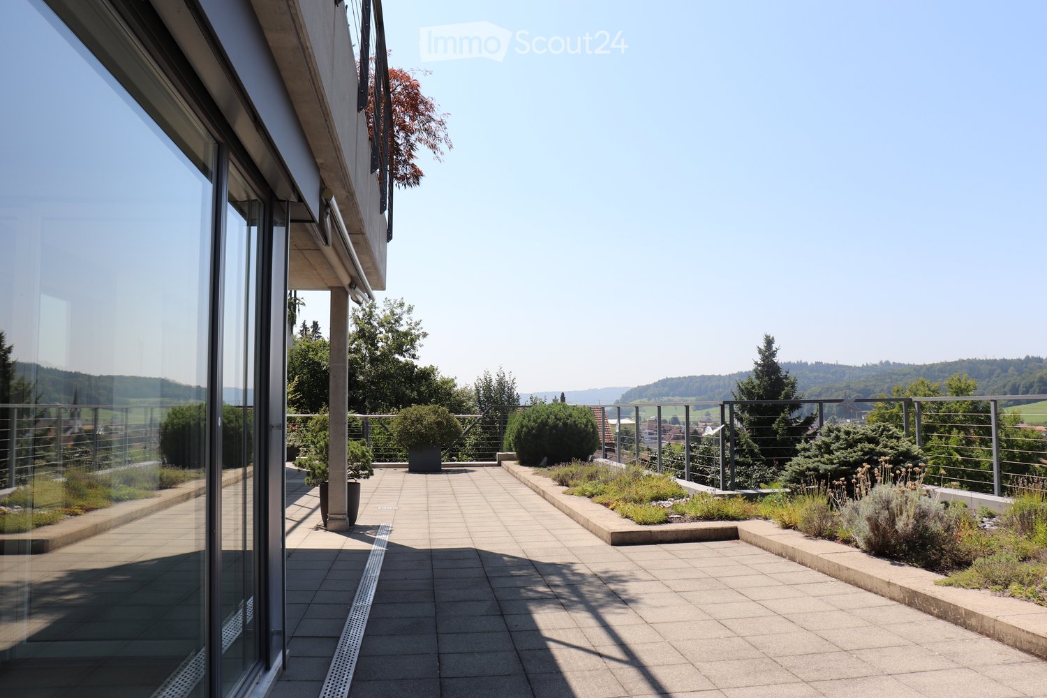 Large outdoor patio with tiled flooring, metal railings, and green plants, with a view of the mountains and cityscape