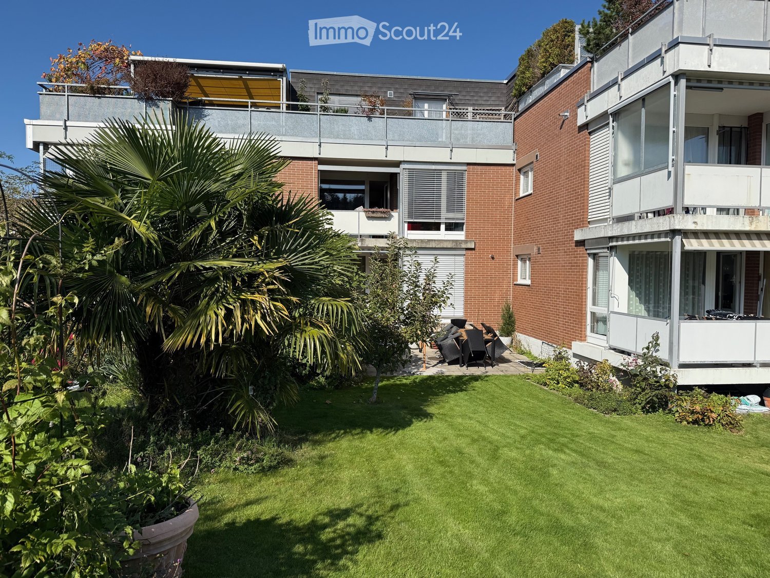 two-story apartment building, brick wall, outdoor terrace, large palm tree, garden with various plants and trees, large green lawn