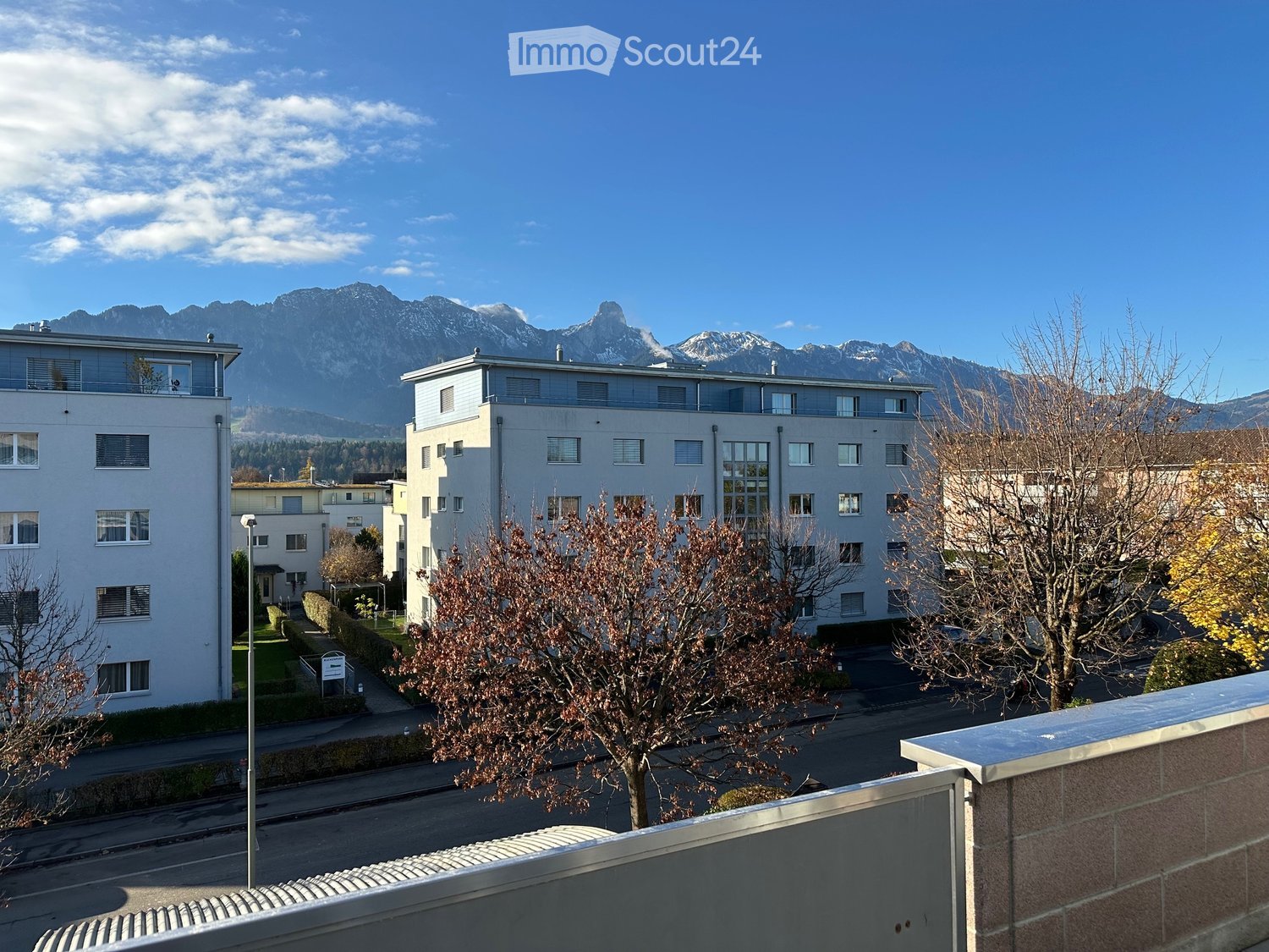 A building with windows facing the mountain, two other buildings visible in the picture, street, sidewalk, trees, blue sky
