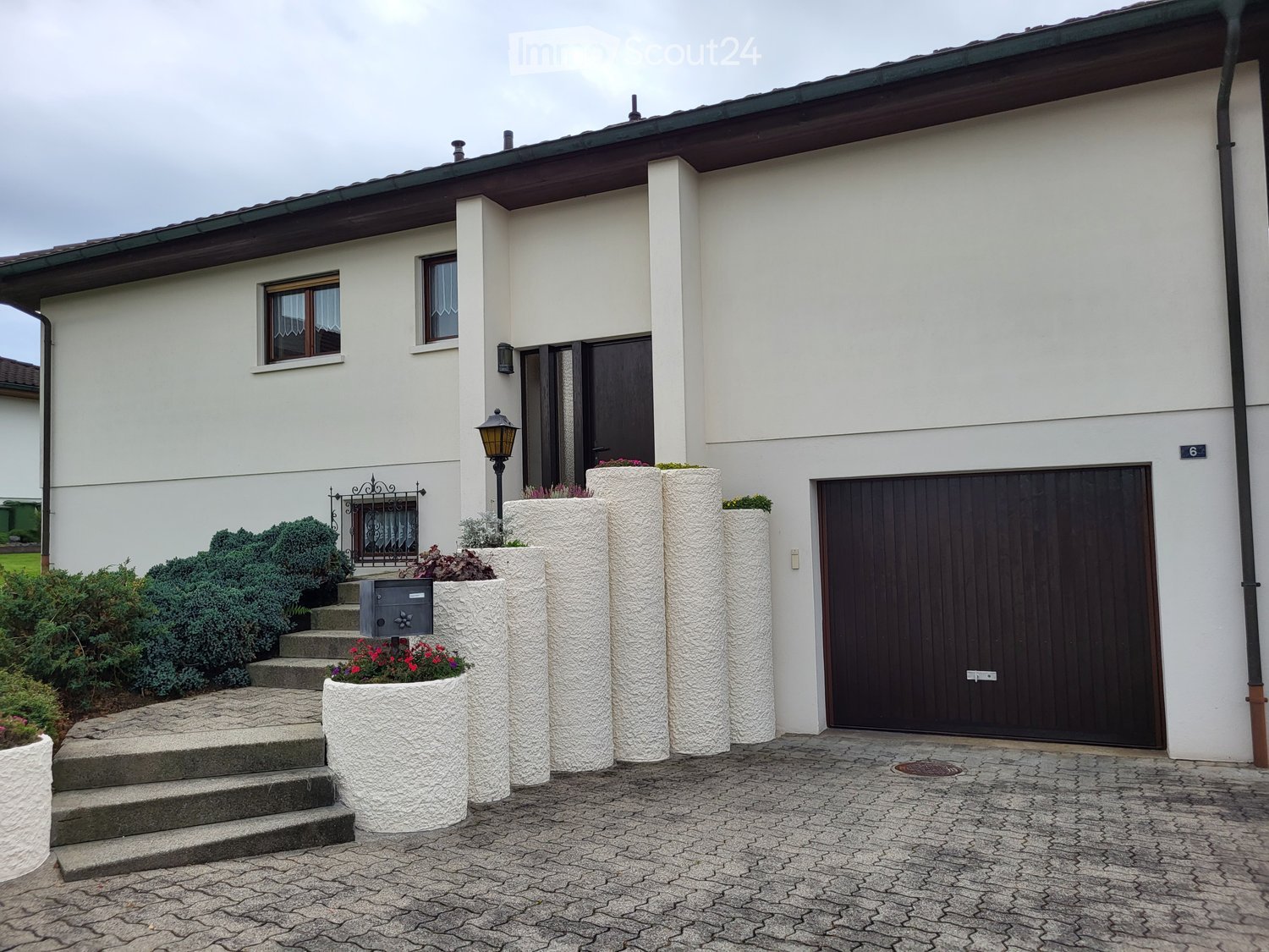 white house, brown garage, large concrete planters with plants, driveway, lamp