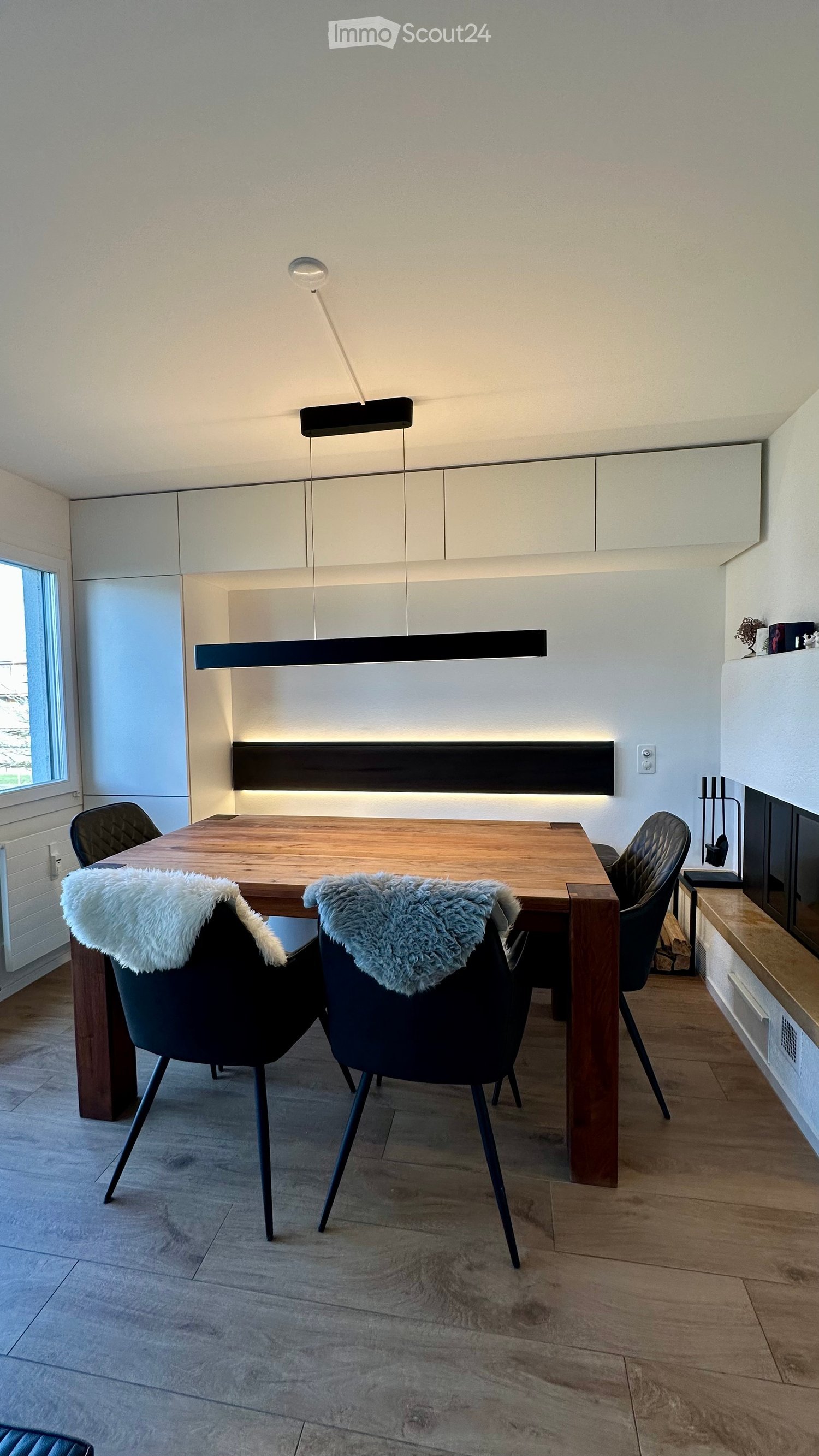 a wooden table with chairs with fur covers, a black pendant lamp, white cupboards, a window and fireplace