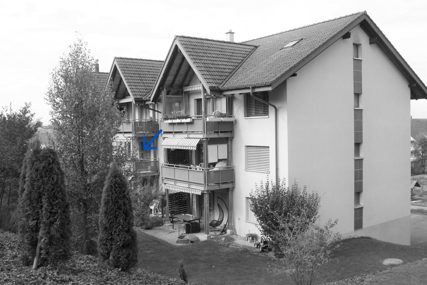 multi-story residential building, gabled roof, balconies, multiple windows, yard with furniture