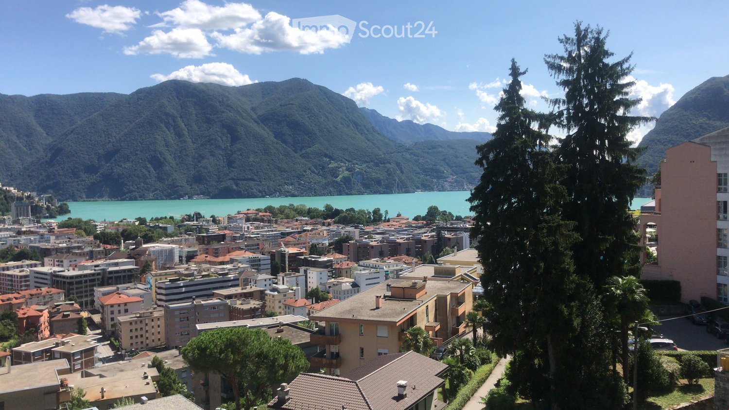Cityscape with multiple buildings of various heights, surrounded by trees, overlooking a lake with mountains in the background.