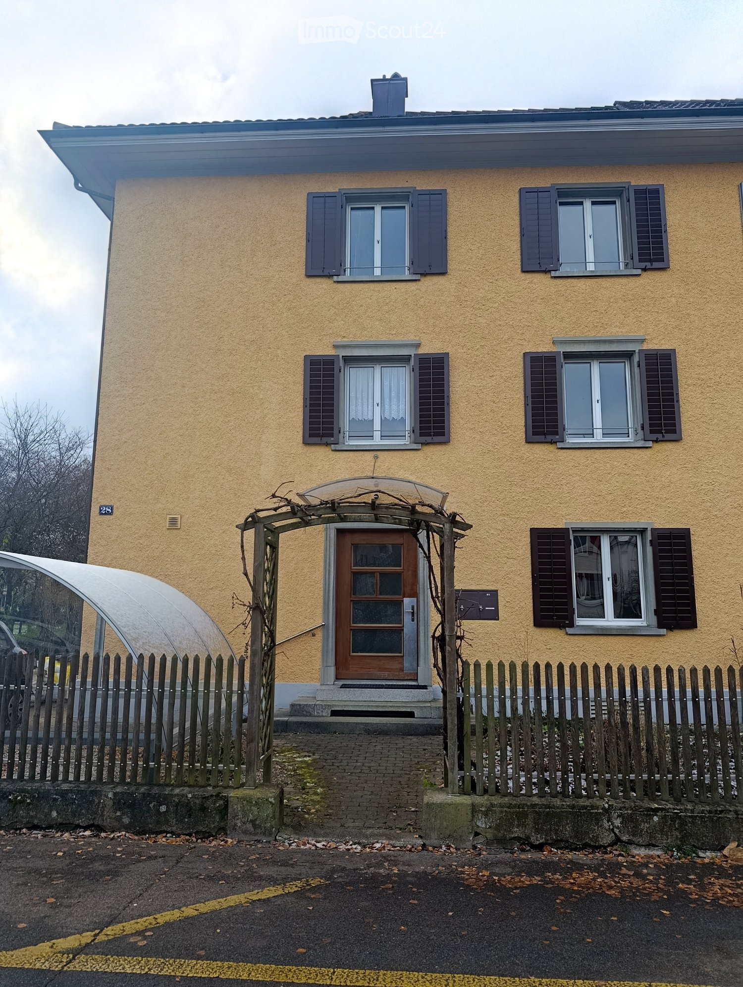 3 story yellow building, brown front door, brown shutters, brick sidewalk, metal fence, parking space