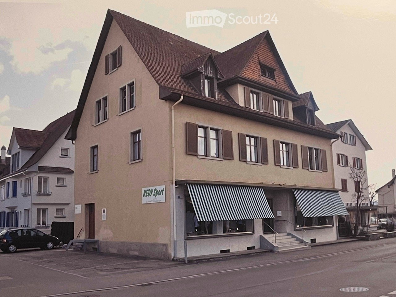 3 story house, brown roof, beige paint, striped awning, steps leading to entrance, parked car in front