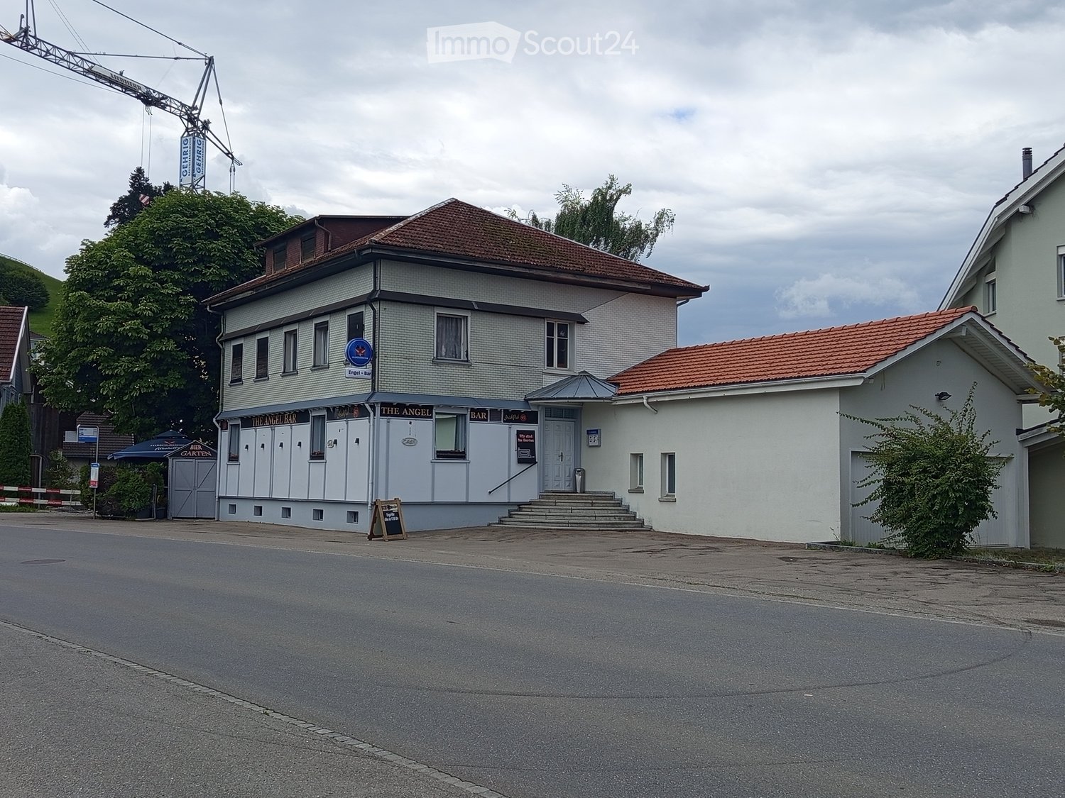 A two-story building with a brown roof and white walls. The entrance is on the ground floor. There is a garden area to the left side with a signboard and plants. A signboard and a blackboard are in front of the building. There is another house on the righ