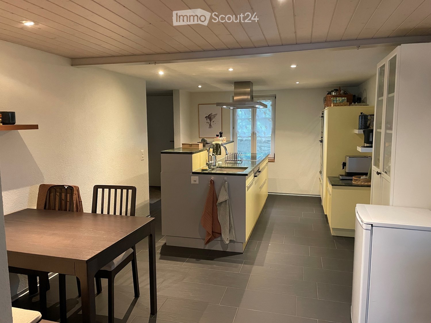 Modern kitchen with island, countertop, faucet, sink, oven, refrigerator, dishwasher, storage cabinets, white tile flooring, open window, lamps, two chairs, wooden table