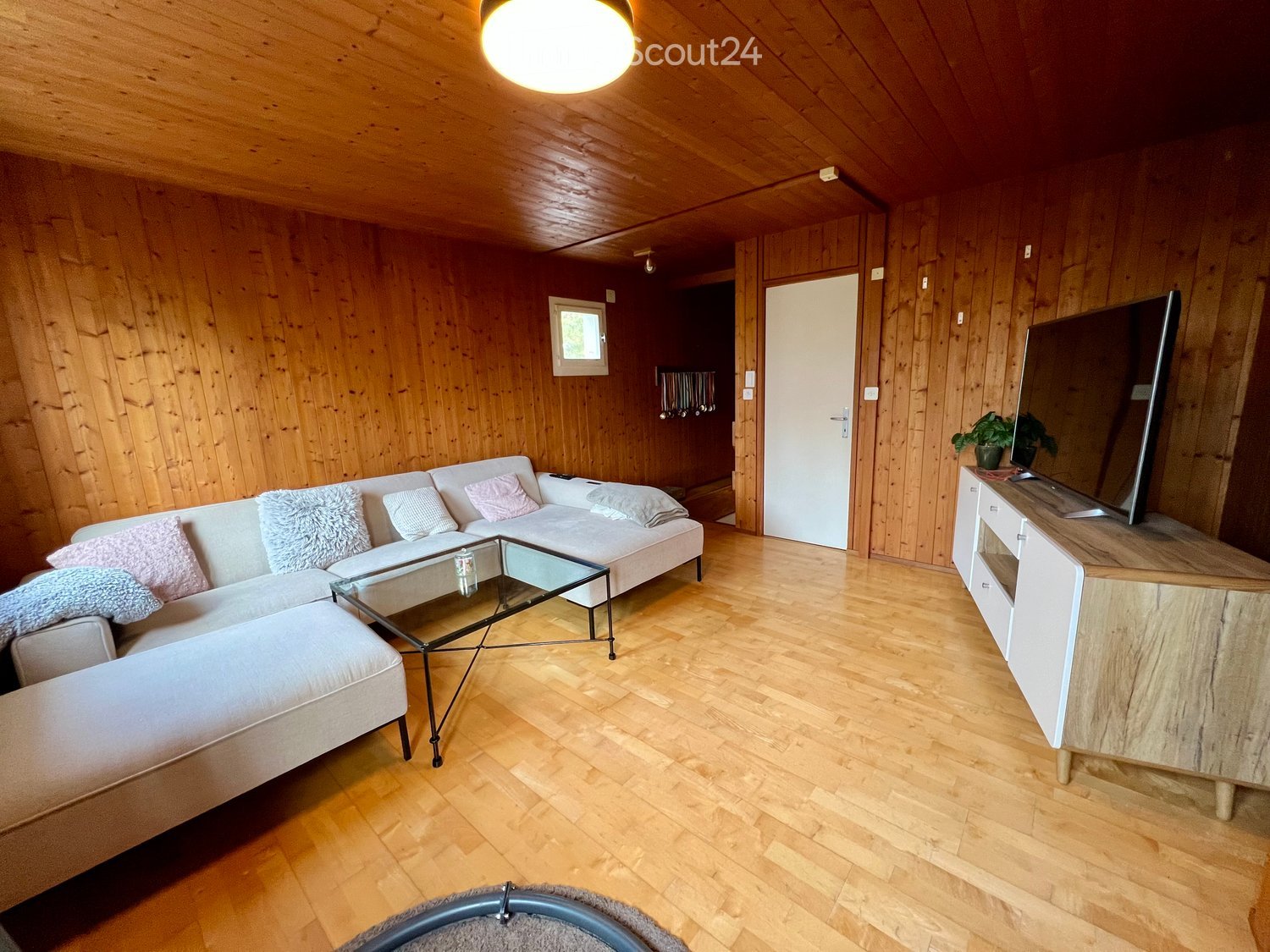 Cozy room with wooden walls, wooden floor, white couch, glass coffee table, TV, potted plant.