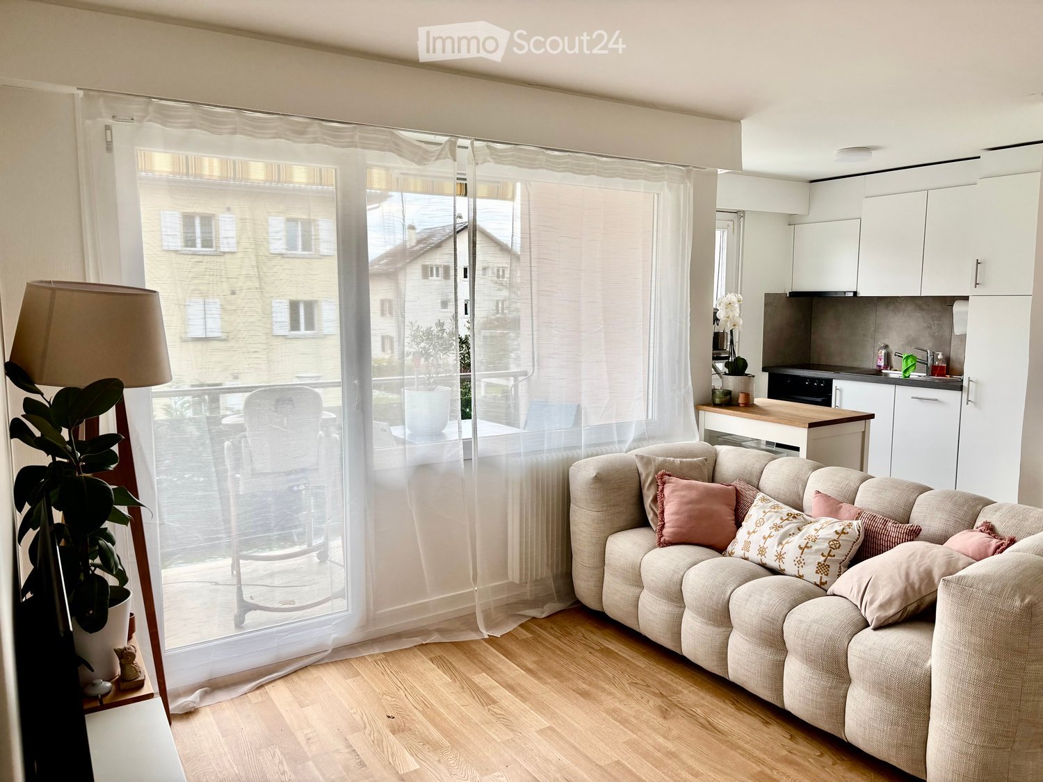 wooden floor, sofa with pillows, large window with curtains, balcony with view of house, white cabinets, kitchen, potted plants