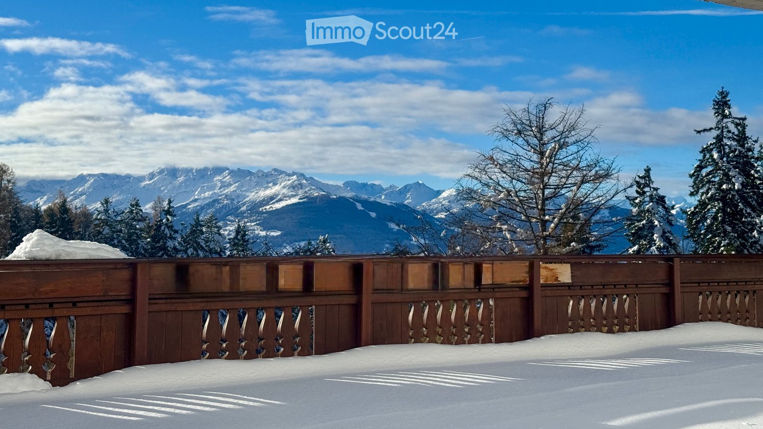 Winter landscape with wooden railing, mountains covered in snow, clear blue sky, pine trees