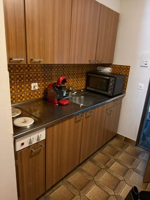 Kitchen area with wooden cupboards, gas stove, sink, microwave, coffee machine, washing machine, tiled floor, power outlets.