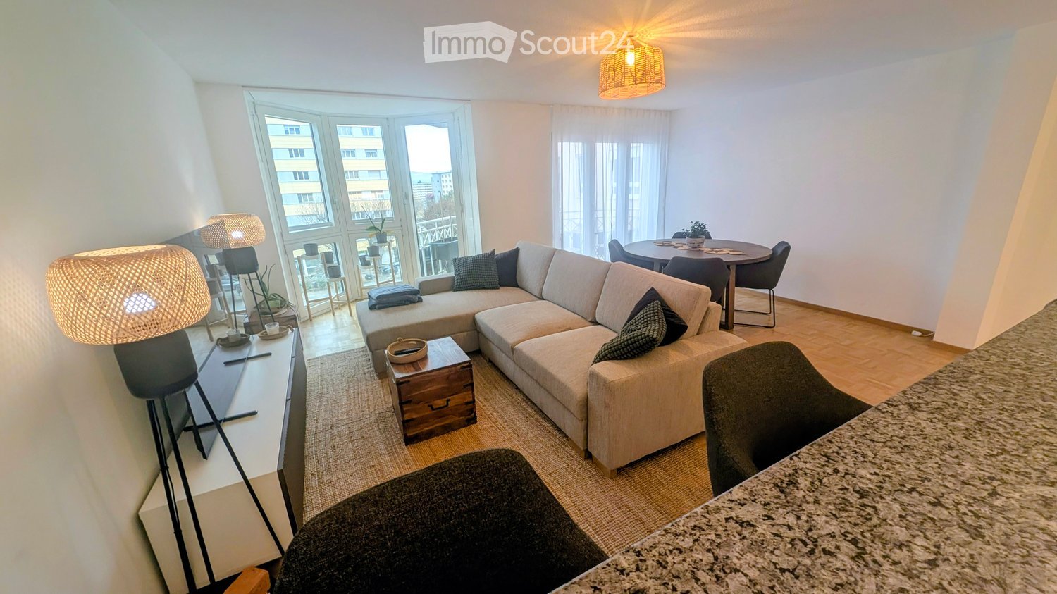 Spacious living room with large windows, L-shaped couch, open kitchen, dining table, lamps, marble counter, carpet