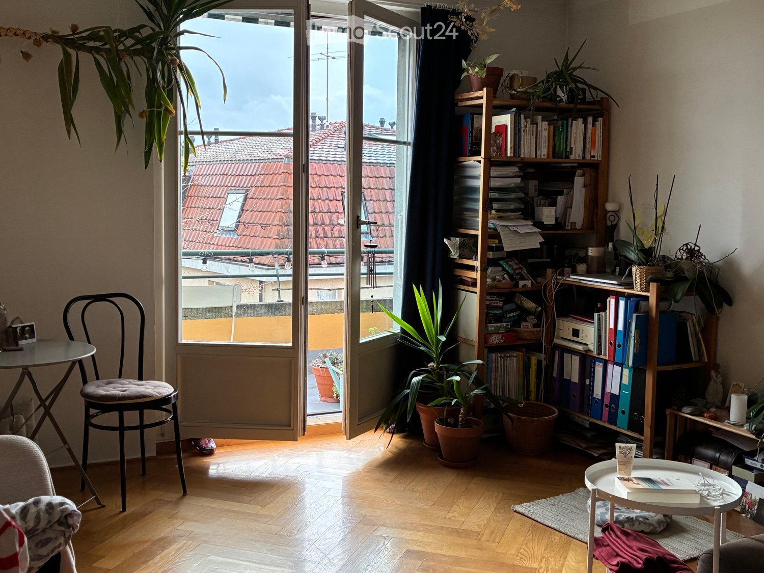 Wooden floors, open doors leading to balcony, bookshelves, plants, furniture, table with book, window