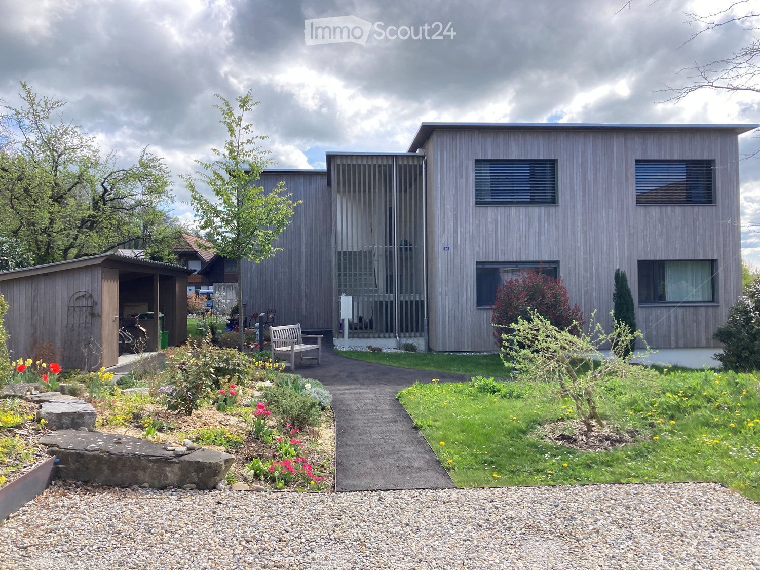 two-story house, wooden exterior, raised ground floor, entrance gate, small garden with plants and a bench