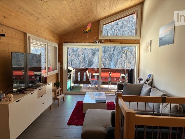 Wood paneling, sliding glass door with balcony, L-shaped couch, two TVs, wooden desk, red rug, coffee table, wooden table and chairs