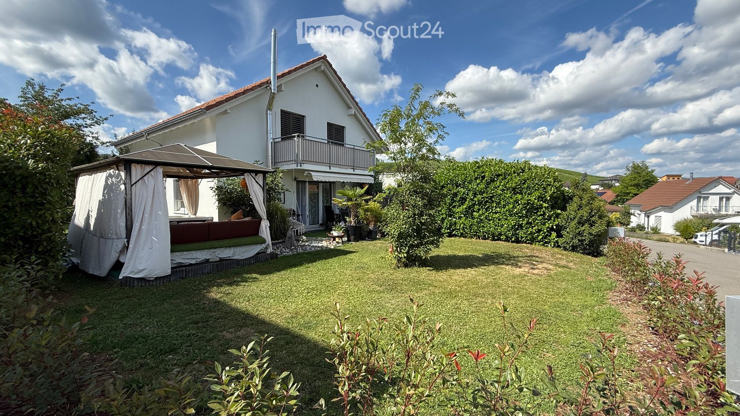 two story house, white paint, brown roof, balcony, patio with canopy, small couch, garden with shrubs and trees
