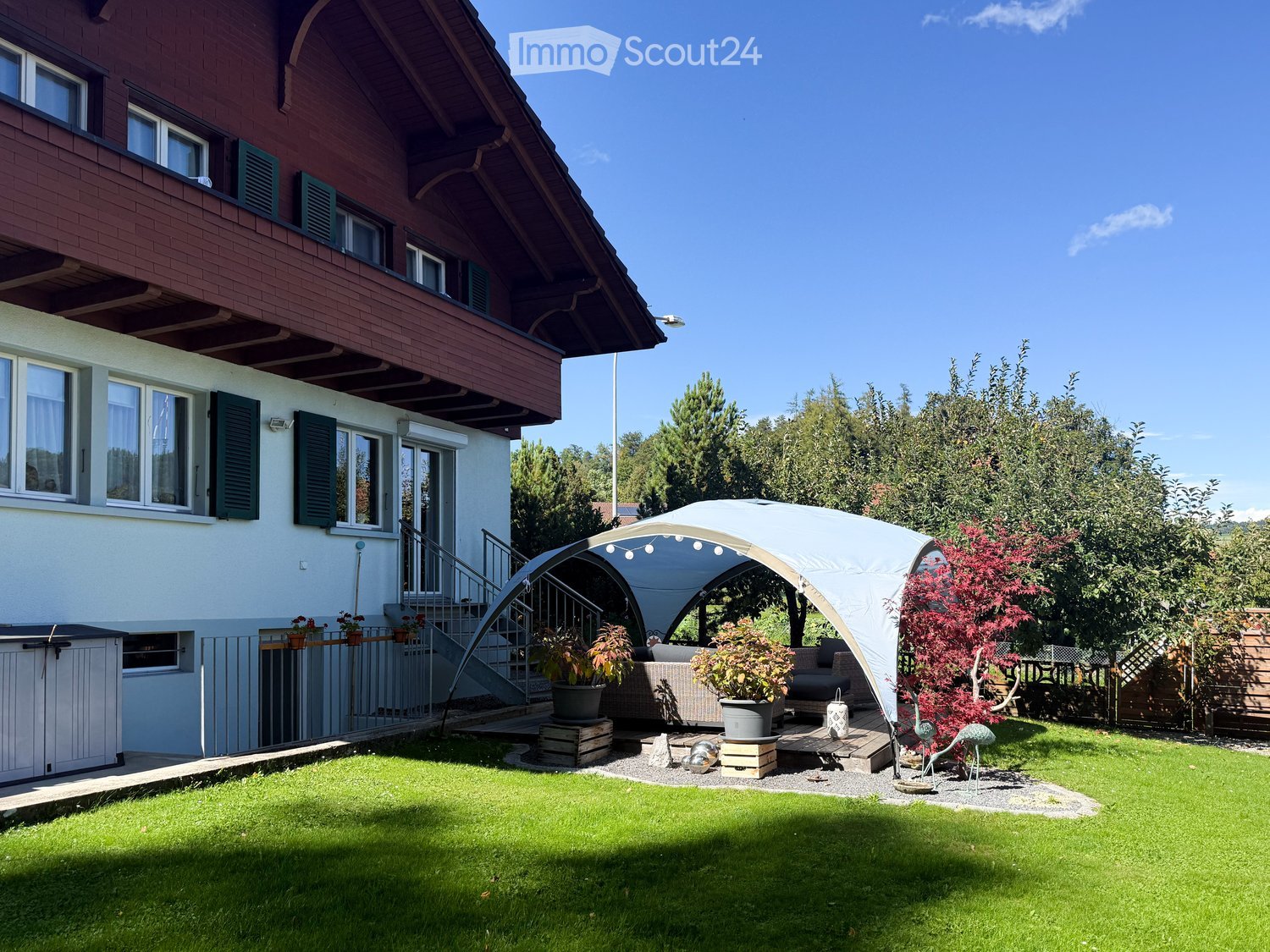 House exterior, brick walls, large windows, brown roof, outdoor tent, couch, potted plants, stairs, grass, trees