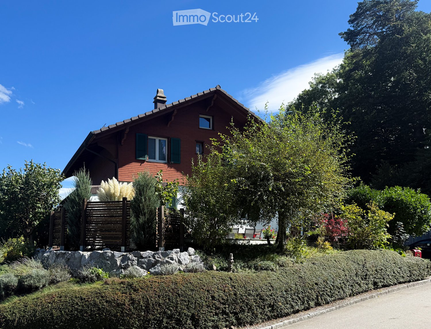 A house with red bricks, green shutters, and a brown roof. The house is surrounded by a garden with various plants and trees.