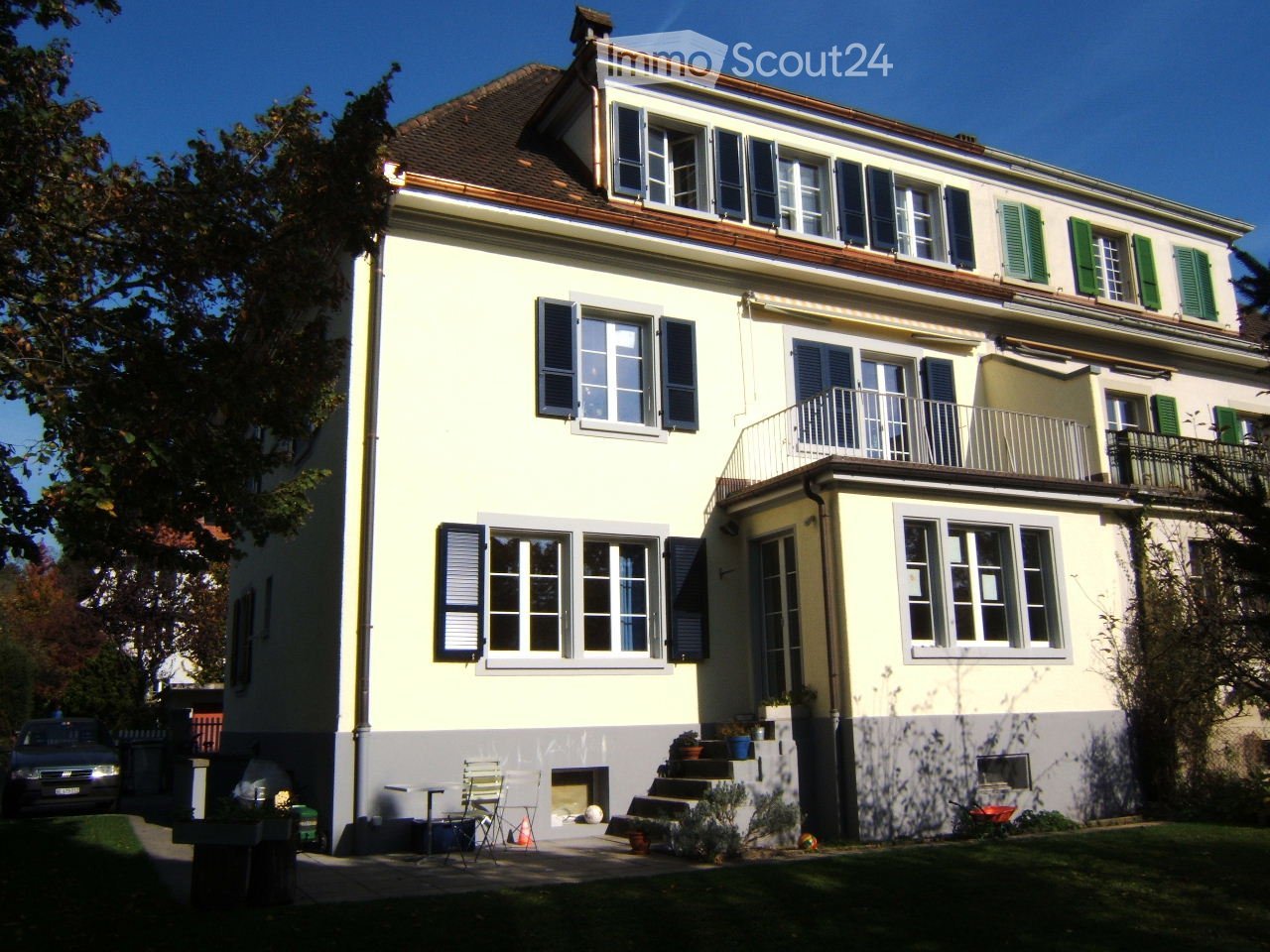 2 story house, yellow exterior, gray lower half, multiple windows, brown roof, balcony, stairs, plants in front