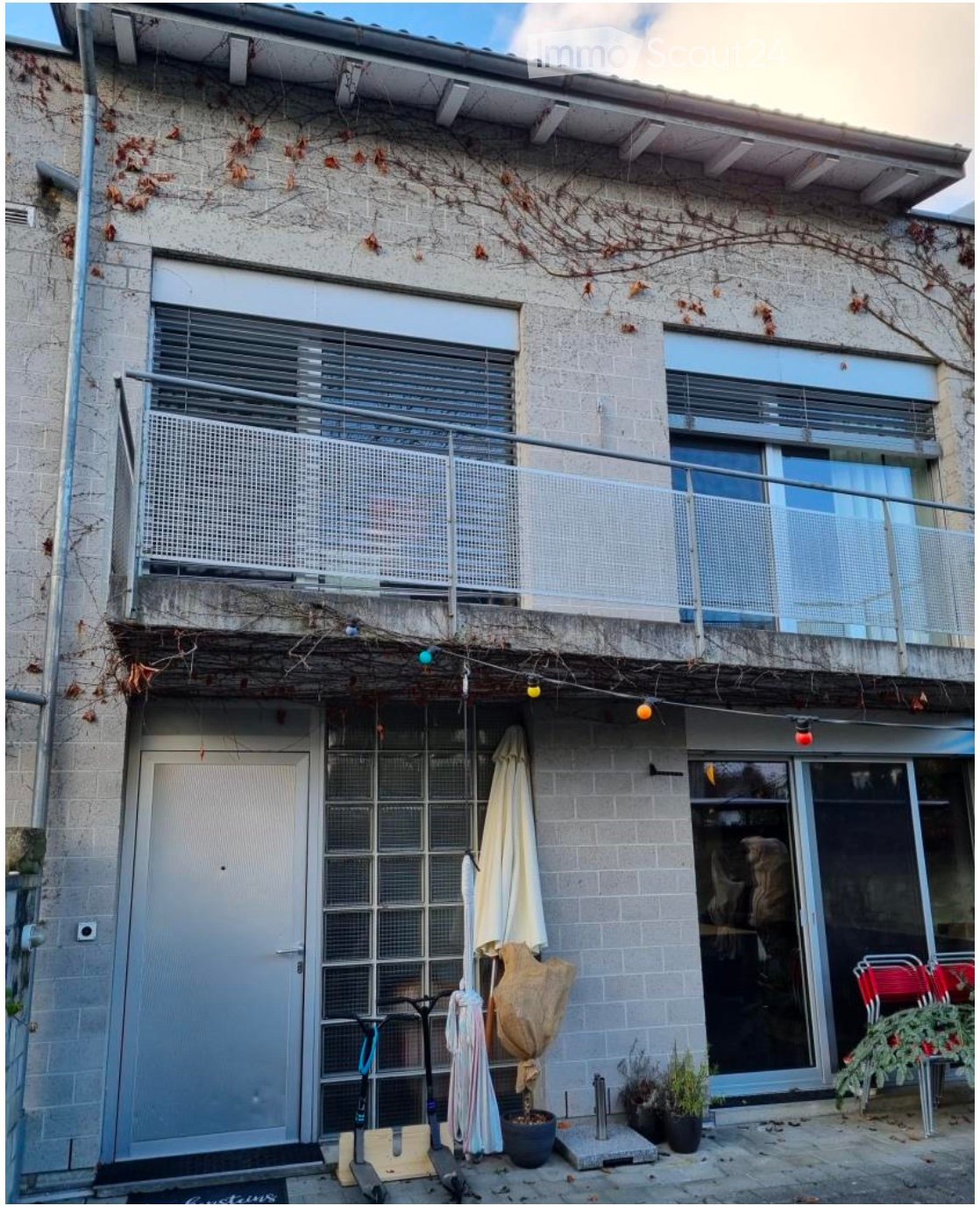 Modern house, brick walls, large balcony with metal railing, entrance with glass paneled door, glass windows, outdoor lighting