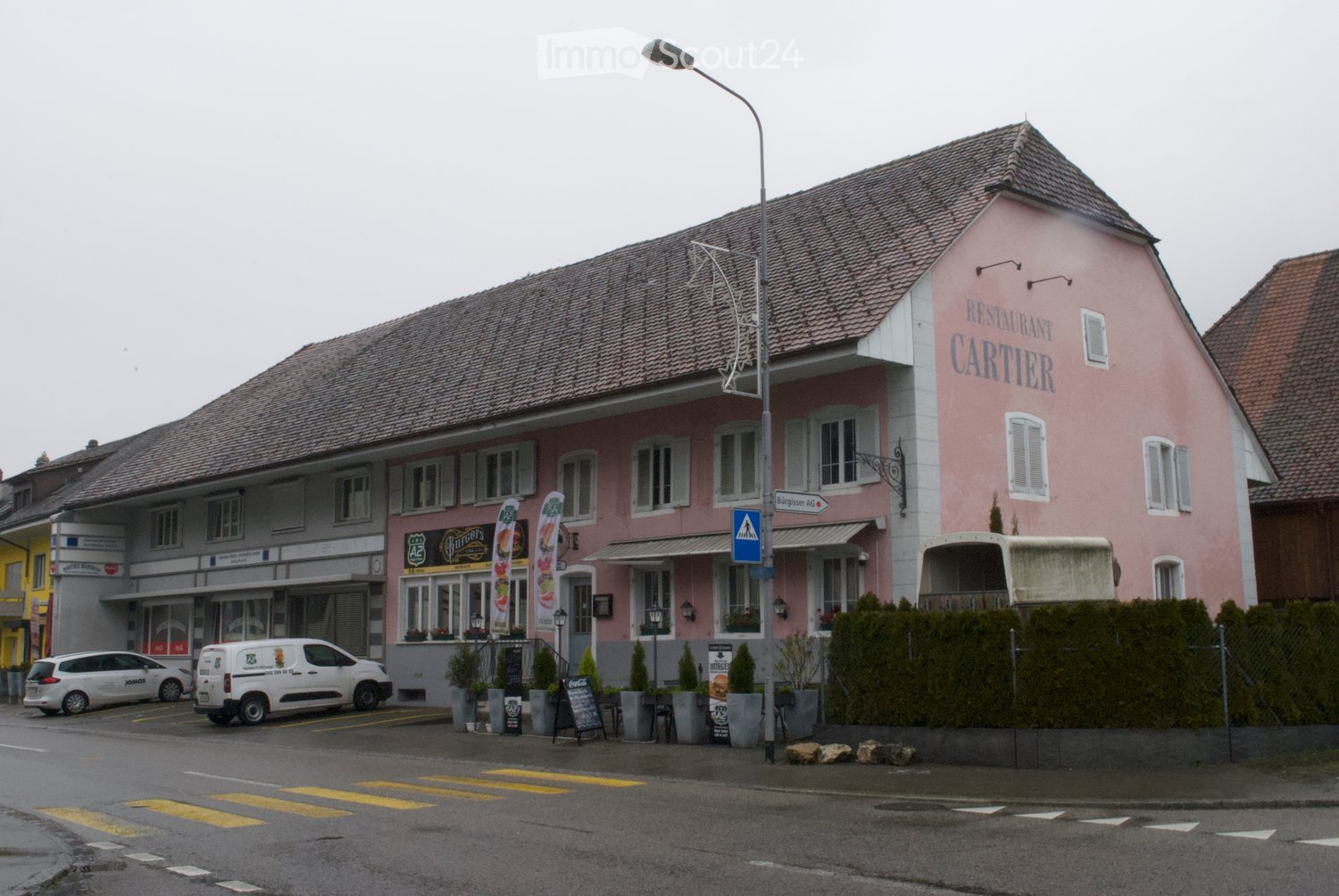 pink painted house, gable roof, sign reading 'RESTAURANT CARTIER', awnings over windows, shrubs, two cars parked in front