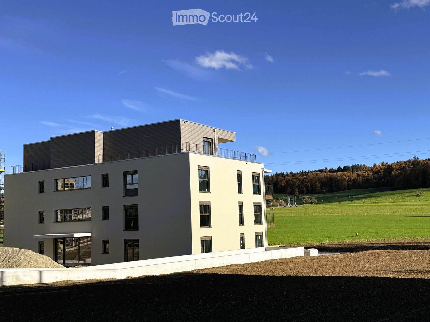 modern white building, multiple floors, large windows, balcony on top floor, green field behind
