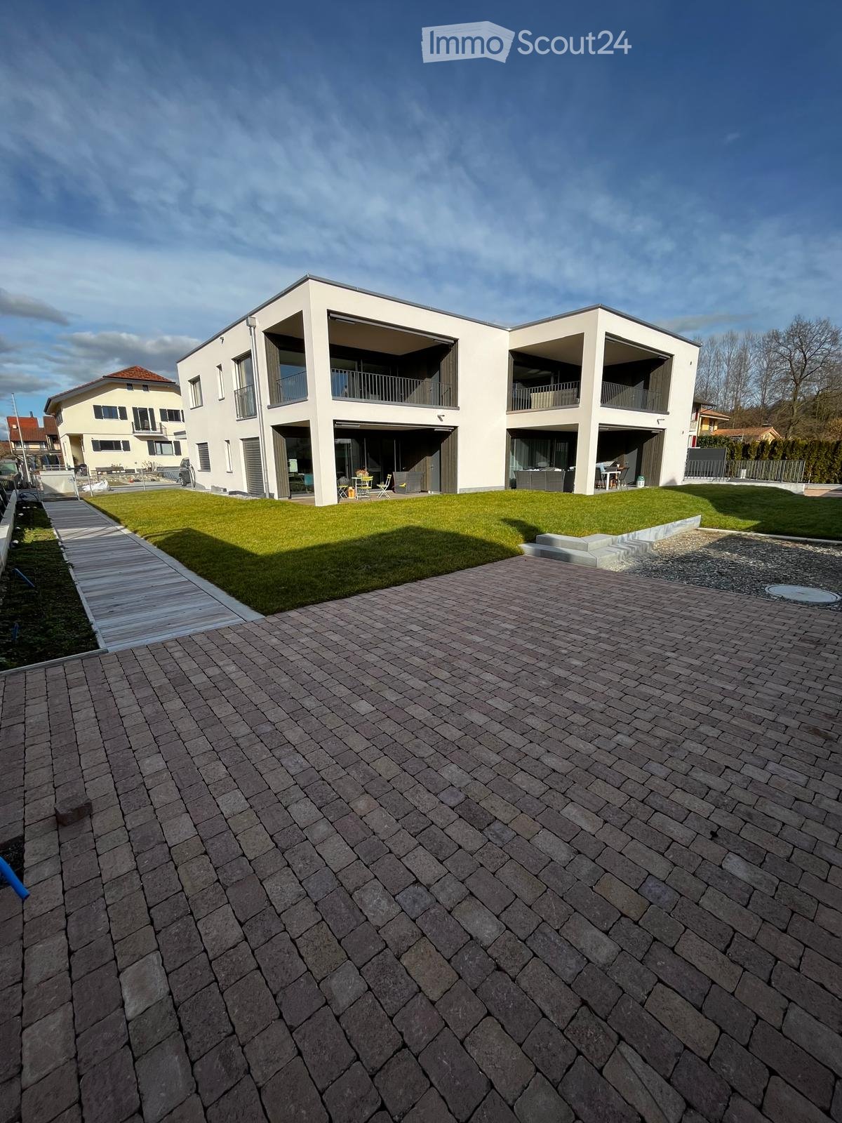 Modern white building with multiple floors, brick driveway, balconies, and well-manicured lawn.