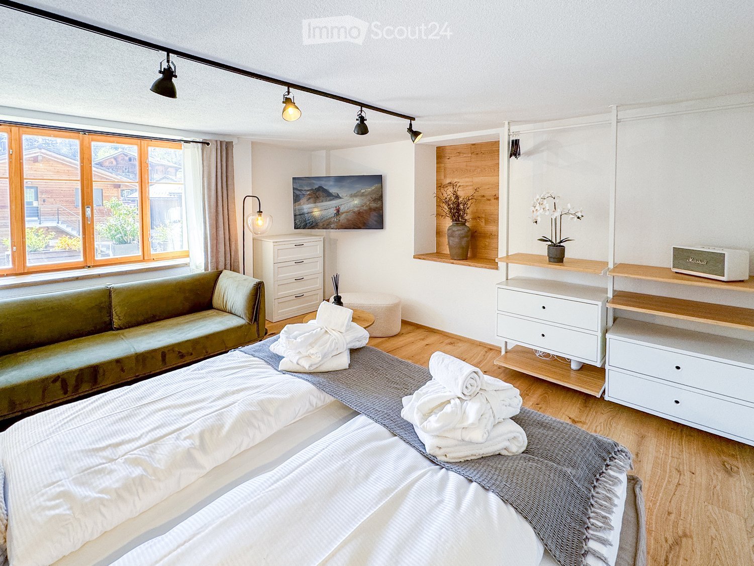 Spacious bedroom with double bed, green couch, TV, wooden floor, white cabinets, bed sheet, towels, shelves, lamps, plants, mounted picture frame