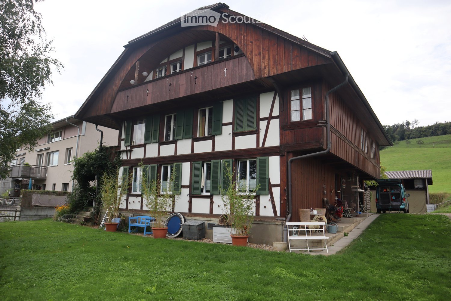 brown wooden house with green shutters, raised ground floor, cars parked at the back, green lawn with potted plants