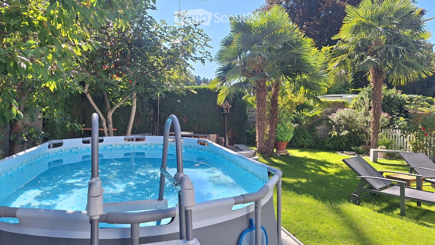 An oval swimming pool is in the garden. A deck chair is on the lawn beside the pool. Plants are around the pool. A bird house is behind the pool. Trees and bushes are in the distance.
