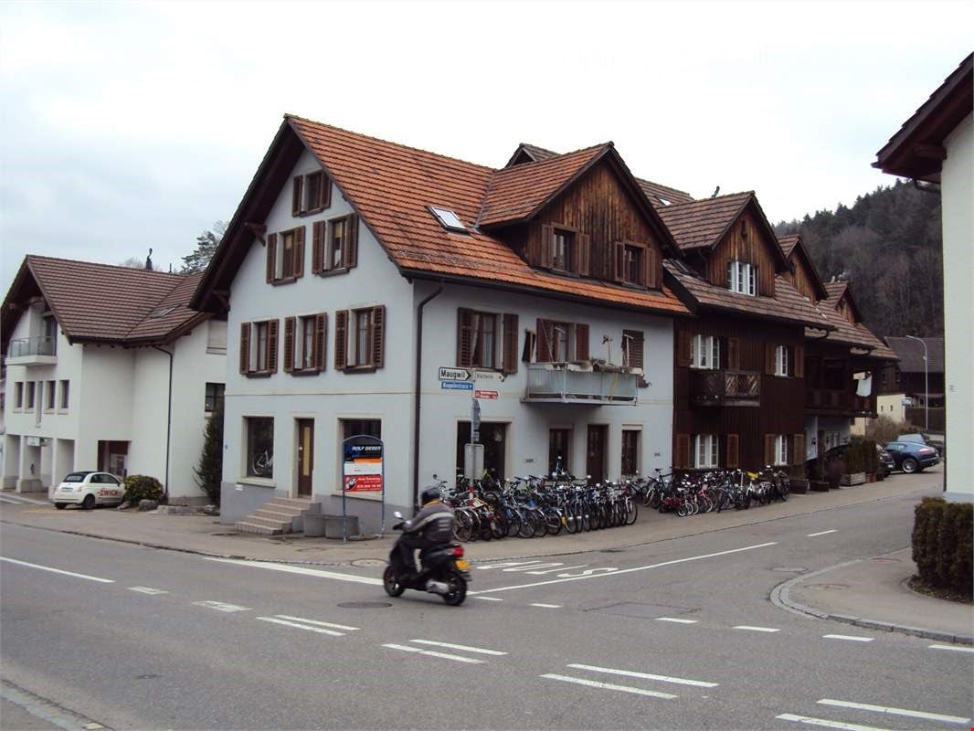 House with red tiled roof, brown wooden facade, white walls, many windows, balconies, cars parked in front, person on scooter