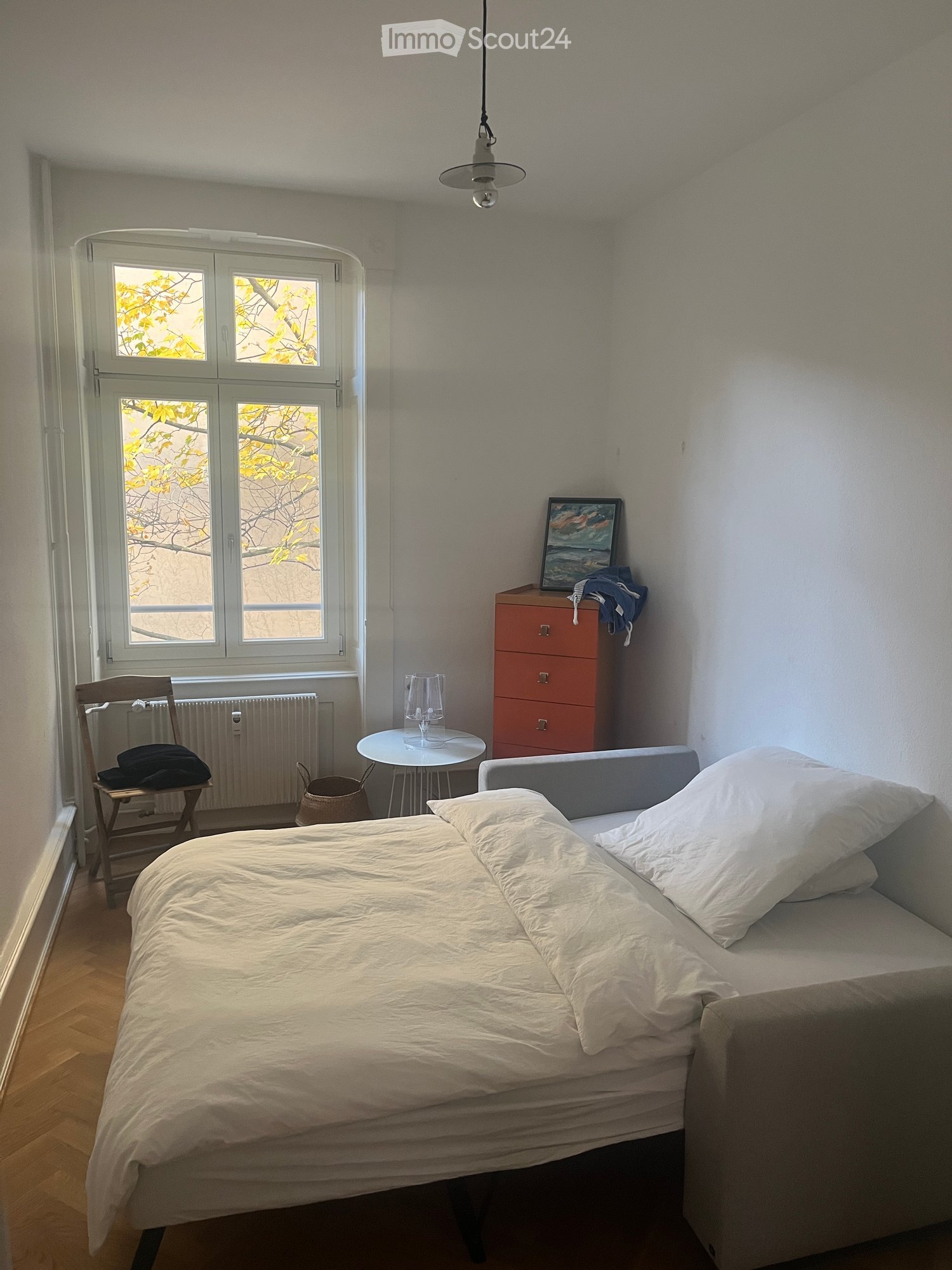 single bed, white walls, wooden floor, window with outside view, wooden chair, small table, cabinet, heater, sofa bed, cushions