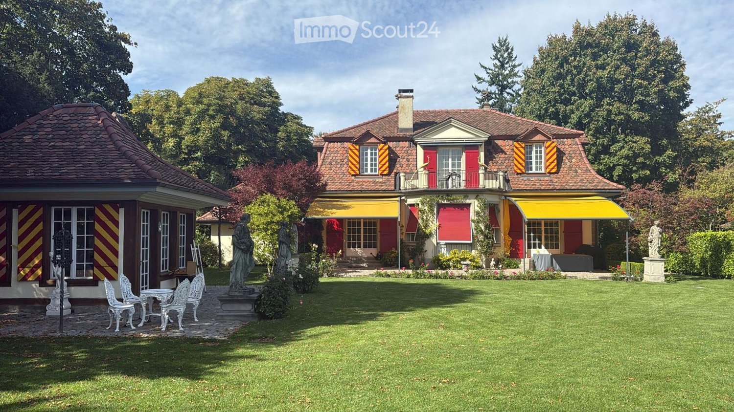 Two houses in the garden, yellow awnings, chimney, green grass, shrubs, plants, two statues, table and chairs in the foreground