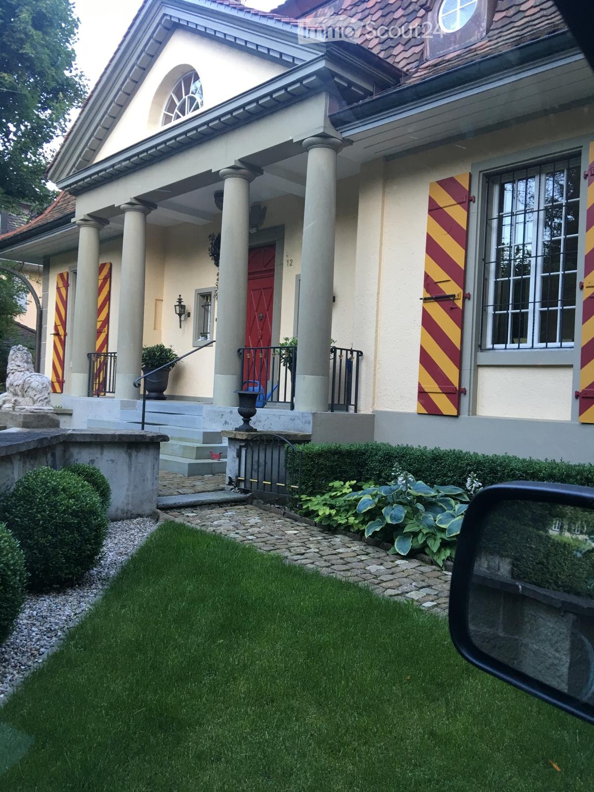 front view of two story house, beige exterior, red door, yellow and red shutters, front garden with lawn, bushes, lion statue, gravel pathway, black iron fence