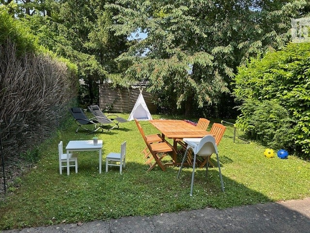 A spacious grassy garden area with wooden table, folding chairs, sun loungers, children's toys, and a teepee tent, all surrounded by a natural setting with trees and shrubs