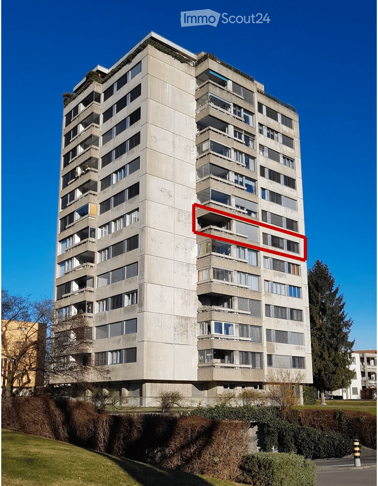 High rise building with many windows, balconies, concrete exterior, green roof, red outline, bush in front
