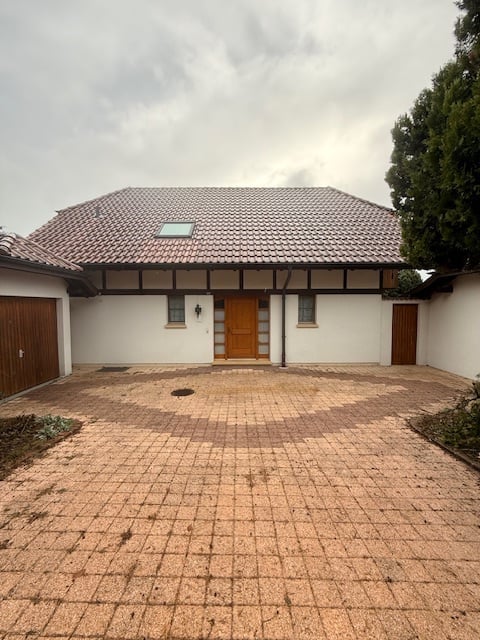 Brown tiled roof, white walls, brown wooden doors, stone paved driveway, two garages, chimney.
